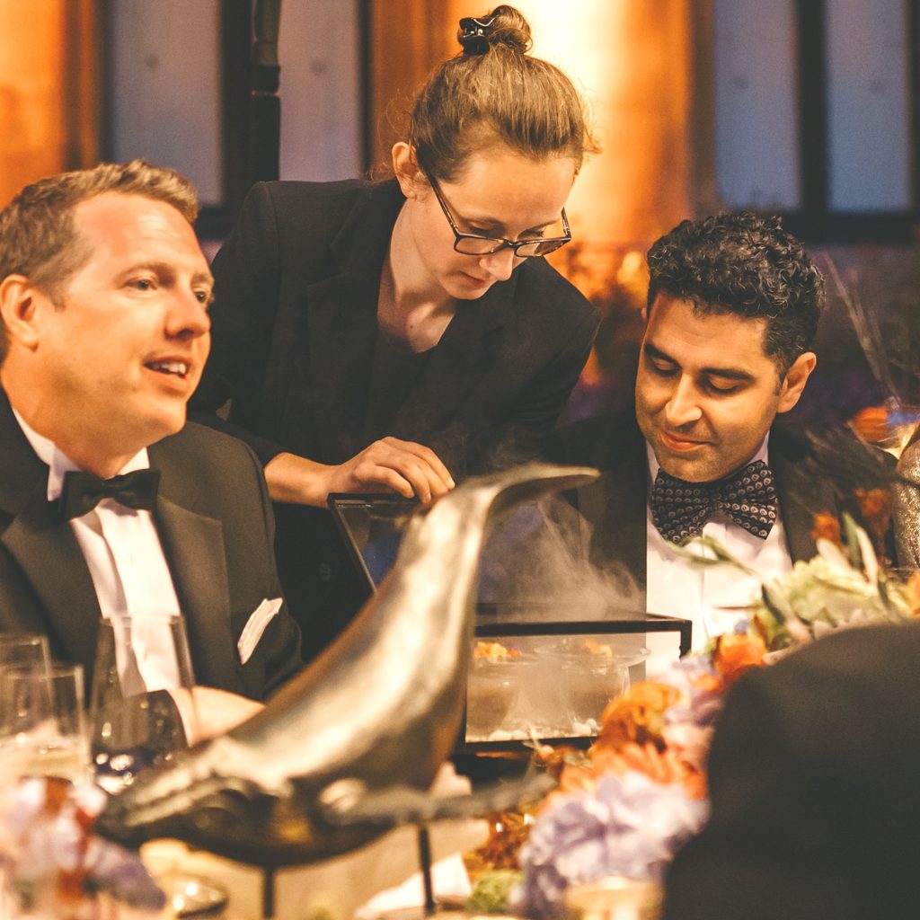 Three people at dinner. One woman opening a box containing food with smoke escaping. A decorative whale placed on the table.