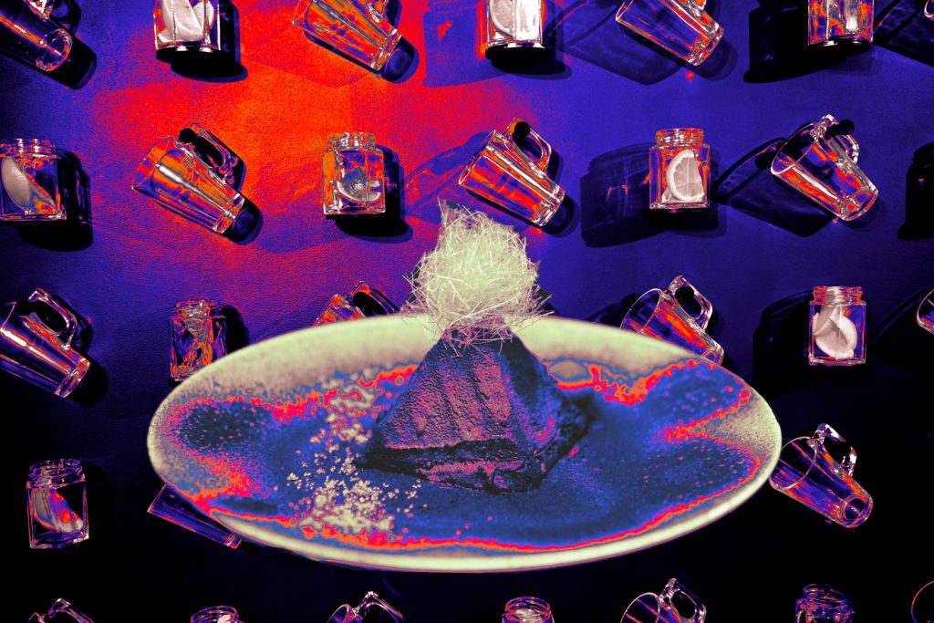 A pyramid shaped dessert on a plate with candy floss sat on top against a purple wall with mounted jars filled with fruit segments.