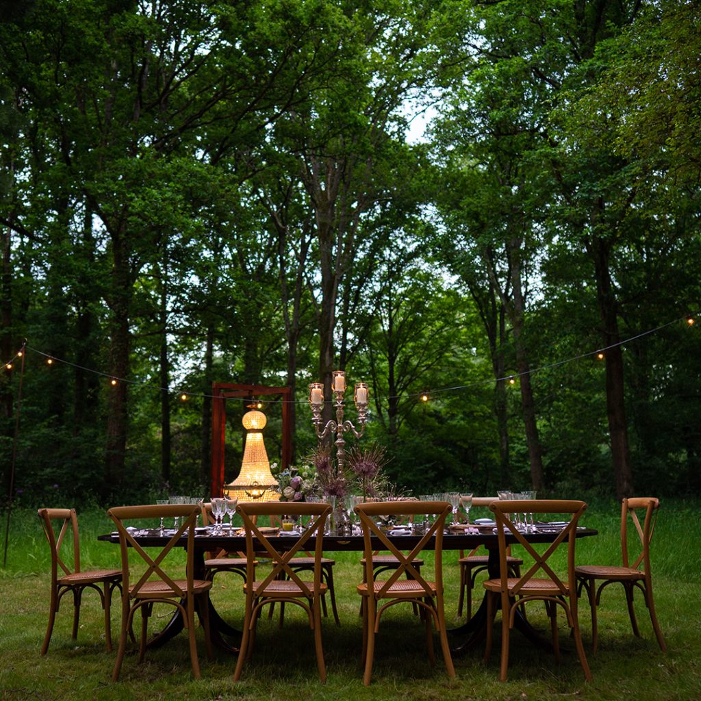 A fine dining wooden dinner table set with mismatched flatware and candles on a lawn in front of a woodlands