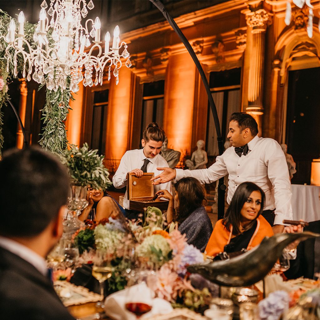Dinner guests under a white chandelier opening a box of chocolate cigars