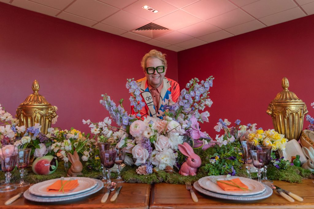A man standing in the middle of a table behind flowers dressed ornately with props, objects and chinaware.