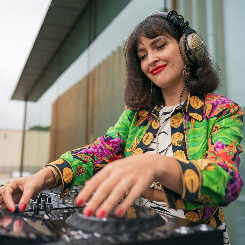 Woman in multi coloured jacked spinning DJ decks wearing headphones