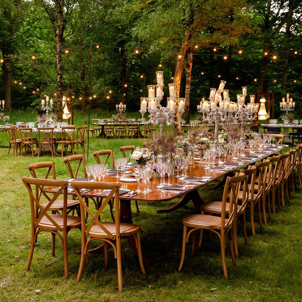 A wooden dining table with wooden dining chairs set on grass with silver candelabra and lit pillar candles