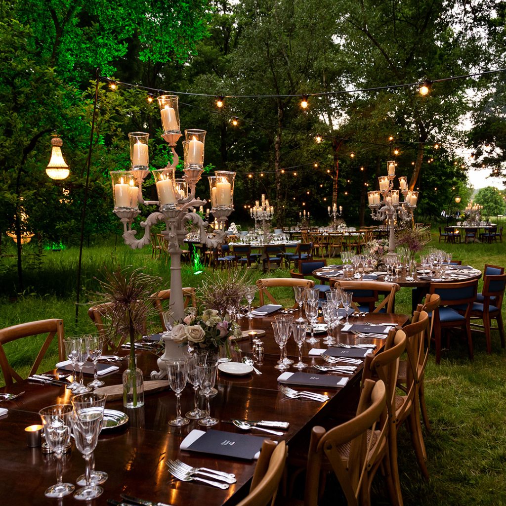 A fine dining wooden dinner table set with mismatched flatware and candles on a lawn in front of a woodlands