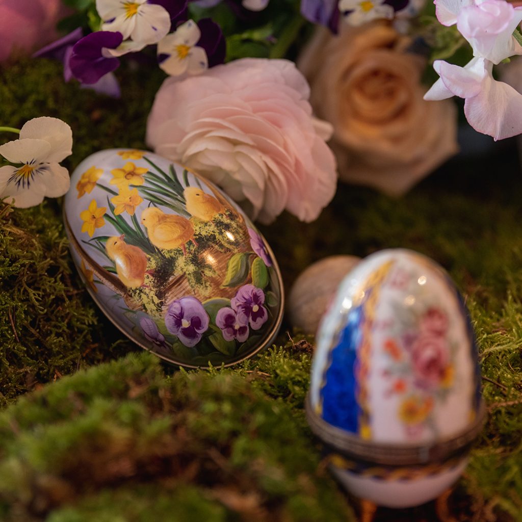 Two decorative china porcelain eggs with metal feet placed on moss and amongst flowers.