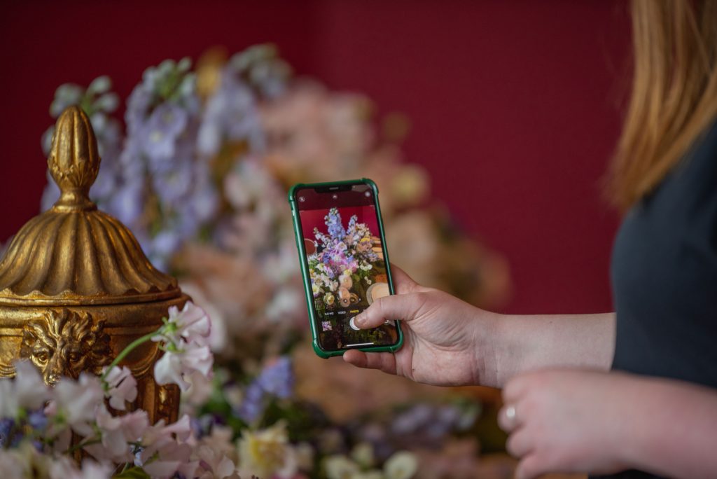 A woman holding a phone taking a picture of flowers and a gold ornate vase