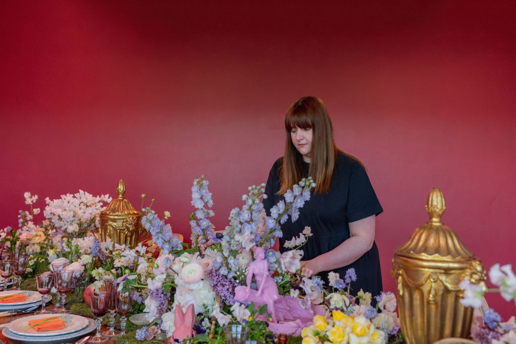 A woman in black attire behind a table of spring flowers and ornate objects in a pink room