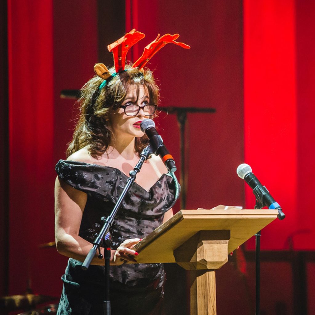 A woman in a black dress at a lectern with red Christmas antler horns on her head.