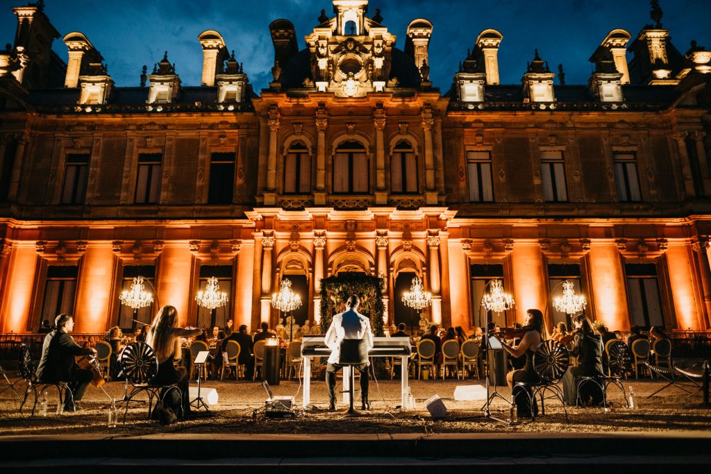 A long table set for dinner in front of Waddesdon Manor. A pianist playing in a white tuxedo. Suspended chandeliers hanging over the table and orange ambient lighting. London event planners.