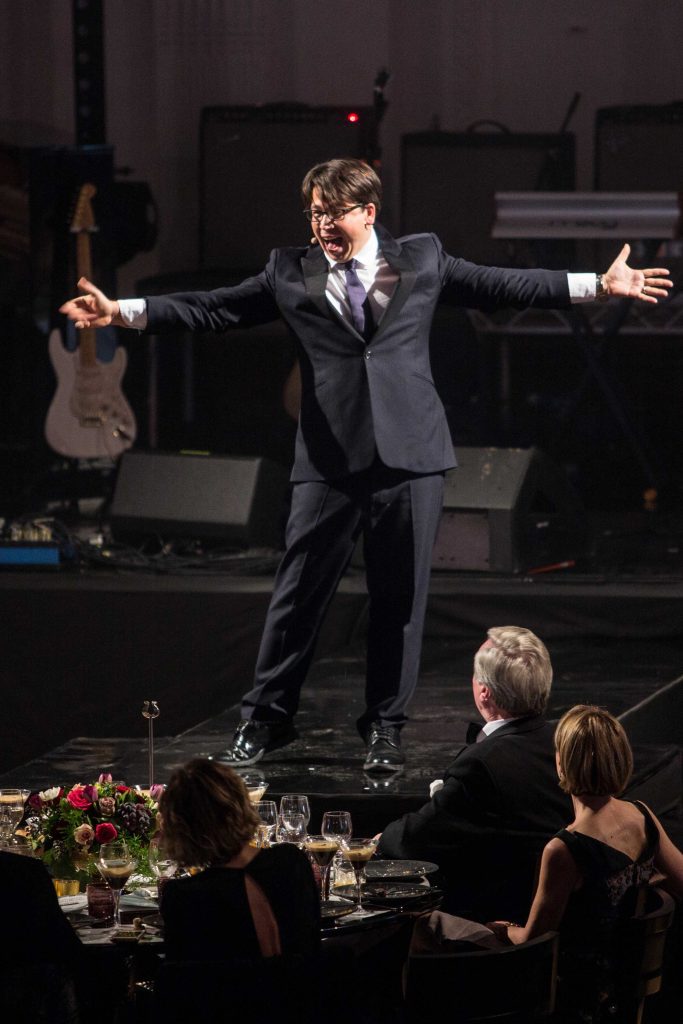 A man in a black suit entertaining a crowd on stage with his arms out wide.