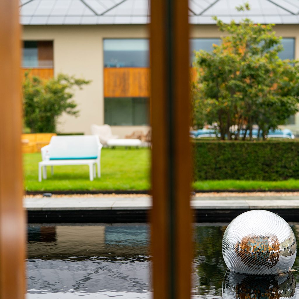 View through a window onto a water feature and lawn. A mirror ball can be seen floating on water and a white chair with blue cushion positioned on the lawn.