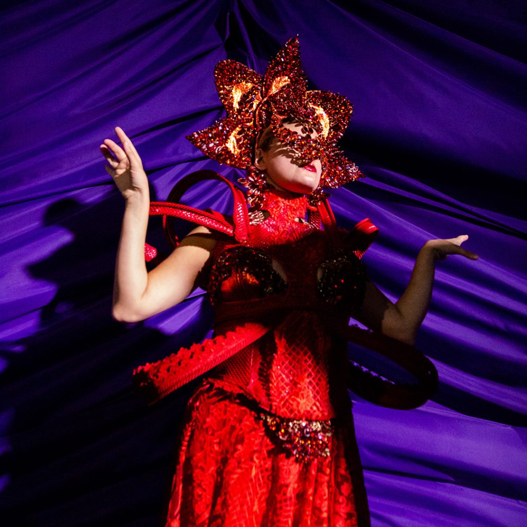 A surrealist performer in red costume in front of a purple drape curtain.