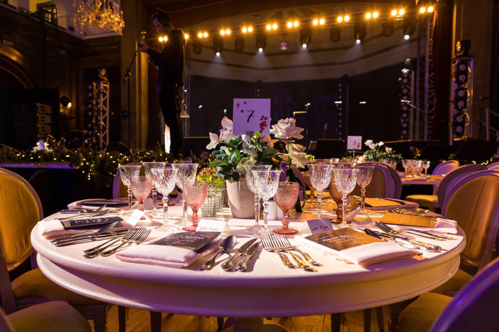 A circular white, wooden dining table with glassware, flowers and flatware set in front of a stage.