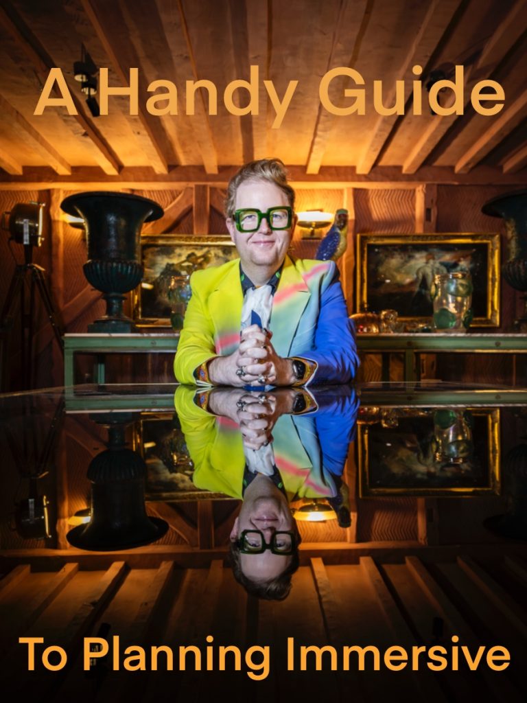 A man sat looking straight onto the camera wearing a blue and yellow jacket with green glasses on a shiny, reflective table.