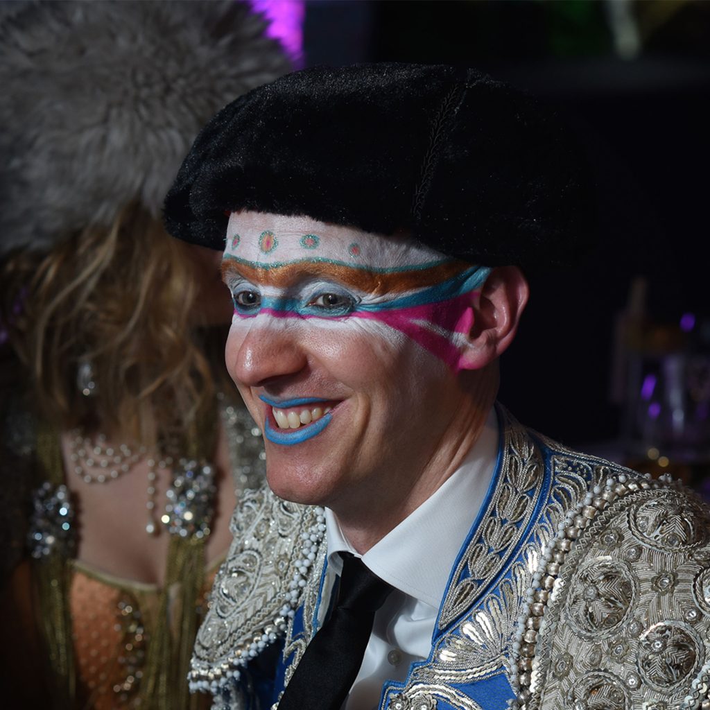 A man wearing an ornate silver suite with a black hat and expressive striped make-up.